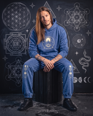 Model 6'0" and 165 lb seated in the royal blue Celestial Synthesis snow wash joggers and matching hand-frayed hoodie, tree of life prints visible on both knees, in front of a chalk sacred geometry wall.