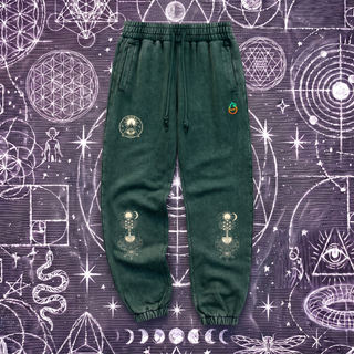 Blackish green Celestial Synthesis snow wash heavyweight jogger sweatpants laid flat on a chalk sacred geometry surface, showing the cream sun and all-seeing eye emblem on the left thigh and the alchemical tree of life on both knees.