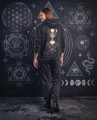 Back view of a model in the black Celestial Synthesis snow wash joggers and hand-frayed hoodie, showing the cream alchemical tree of life print on the hoodie back, in front of a chalk sacred geometry wall.