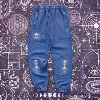 Royal blue Celestial Synthesis snow wash heavyweight jogger sweatpants laid flat on a chalk sacred geometry surface, showing the cream sun and all-seeing eye emblem on the left thigh and the alchemical tree of life on both knees.