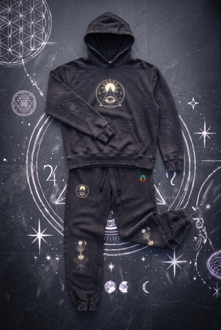 Black Celestial Synthesis hoodie and joggers set laid out as one outfit on dark slate with white chalk sacred geometry, cream sun and all-seeing eye emblem on the hoodie chest and jogger thigh, tree of life on both knees.
