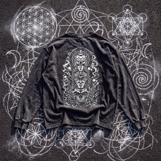 Back of the Paradigm acid wash oversized long sleeve tee in Black Beauty, styled on dark asphalt covered in white chalk sacred geometry, showing a tall arched panel of stacked ornate faces wrapped in smoke and orbs.