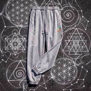 Paradigm heavyweight snow wash joggers in cool Light Gray, front view, laid on dark asphalt covered in white chalk sacred geometry, showing the white impossible-geometry cube print on the thigh, the pH logo on the hip, the drawcord waistband and ribbed ankle cuffs.