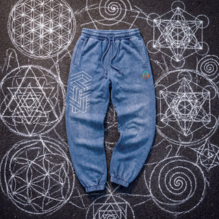 Paradigm heavyweight snow wash joggers in washed Royal Blue, front view, laid on dark asphalt covered in white chalk sacred geometry, showing the white impossible-geometry cube print on the thigh, the pH logo on the hip, the drawcord waistband and ribbed ankle cuffs.