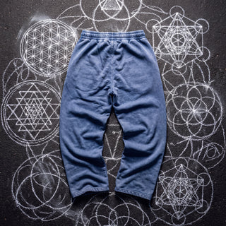 Back of the Paradigm snow washed baggy heavyweight sweatpants in dark blue, laid flat on dark asphalt covered in white chalk sacred geometry, showing the plain back, elastic waistband and wide straight legs.