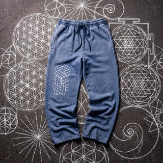 Front of the Paradigm snow washed baggy heavyweight sweatpants in dark blue, laid flat on dark asphalt covered in white chalk sacred geometry, with a white impossible-geometry column of interlocking beams printed on the right thigh, a drawcord waistband and wide straight legs.