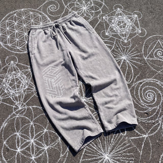 Front of the Paradigm snow washed baggy heavyweight sweatpants in light gray, laid flat on dark asphalt covered in white chalk sacred geometry, with a white impossible-geometry column of interlocking beams printed on the right thigh, a drawcord waistband and wide straight legs.