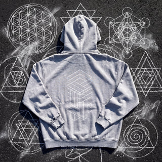 Back of the Paradigm snow wash hand-frayed heavyweight hoodie in light gray, laid flat on dark asphalt covered in white chalk sacred geometry, printed with a tall white impossible-geometry column of interlocking beams.