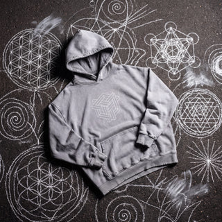 Front of the Paradigm snow wash hand-frayed heavyweight hoodie in light gray, laid flat on dark asphalt covered in white chalk sacred geometry, with a white impossible-geometry cube knot printed on the chest, a kangaroo pocket and a hand-frayed hood, cuffs and hem.