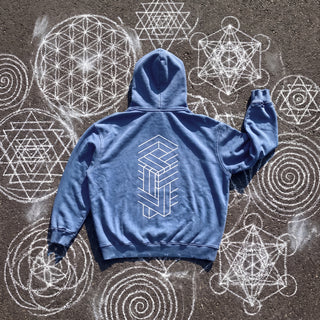 Back of the Paradigm snow wash hand-frayed heavyweight hoodie in royal blue, laid flat on dark asphalt covered in white chalk sacred geometry, printed with a tall white impossible-geometry column of interlocking beams.