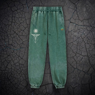 Green acid-wash sweatpants with a white geometric sun-and-pentagram graphic on the left leg and a small multicolor circular graphic on the right leg, displayed flat against a cracked dark gray ground background.