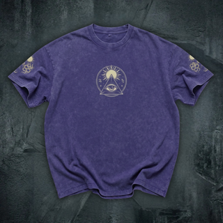 A purple short-sleeve t-shirt with a central gold-toned all-seeing eye and sun graphic, laid flat on a dark textured background.