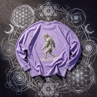 Back of the PsySquatch acid wash oversized long sleeve tee in Purple, styled on dark asphalt covered in white chalk sacred geometry, showing a bigfoot walking in front of a flower of life halo.
