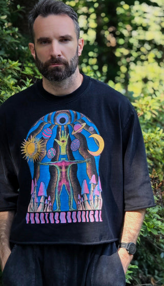 A man wearing a black graphic t-shirt with a colorful psychedelic design, standing outdoors in front of a blurred green foliage background.