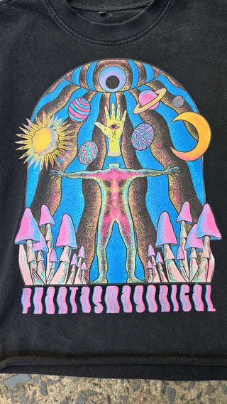 A close-up of a black T-shirt featuring a large psychedelic graphic print with a stylized face, crescent moon, sun, abstract body, and pink striped vertical text on a black background.