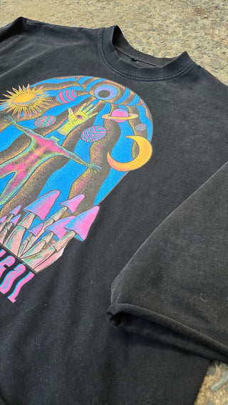 A close-up of a black graphic t-shirt laid flat on a textured concrete surface. The shirt features a large, colorful psychedelic print on the front depicting a stylized figure with a crescent moon, sun, and pink mushroom shapes.