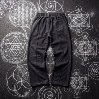 Back of the Spirit Molecule snow wash drawstring cotton pants in black, laid flat on dark asphalt covered in white chalk sacred geometry, showing the elastic waistband, a single back patch pocket and the wide baggy legs.