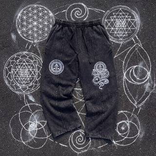 Front of the Spirit Molecule snow wash drawstring cotton pants in black, laid flat on dark asphalt covered in white chalk sacred geometry, with a white circular meditation seal printed on one thigh and a white centipede serpent print running down the other leg.
