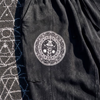 Close-up of the Spirit Molecule pants thigh artwork: a white circular seal with a sacred geometry border, a ring of moon phases, a meditating figure, a stack of three eyes and two orbs, on washed black cotton.