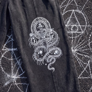 Close-up of the Spirit Molecule pants serpent artwork: a small circular seal above a fanged centipede serpent coiling down the leg among floating orbs, in white on washed black cotton.