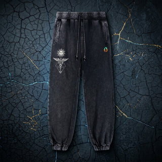 Black snow wash Inner Alchemy 420 GSM joggers laid flat on cracked dark stone, with the cream sun and winged caduceus thigh print and the pH flame logo on the hip